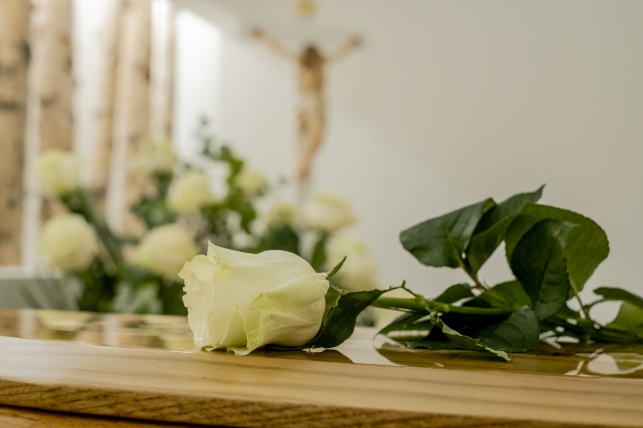 Primo piano di una rosa bianca con un crocifisso sfocato sullo sfondo durante un rito funebre