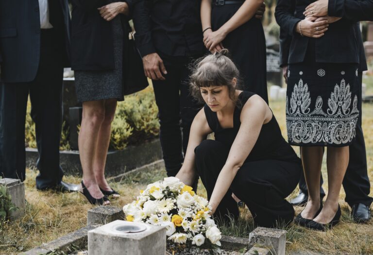 Morte apparente: quanto può durare? Donna al cimitero inginocchiata e con i familiari alle sue spalle, mentre appoggia un mazzo di fiori bianchi sulla tomba di una persona cara defunta